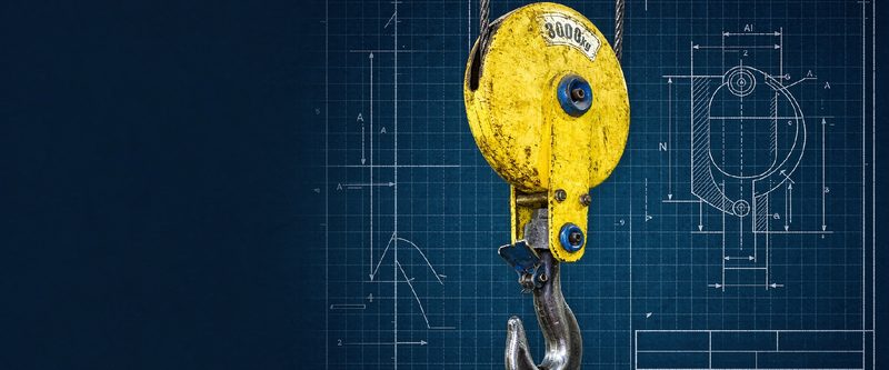 Steel pulley block with hook over an engineering blueprint marked DIN, ISO, EN, ASTM, GB and JIS